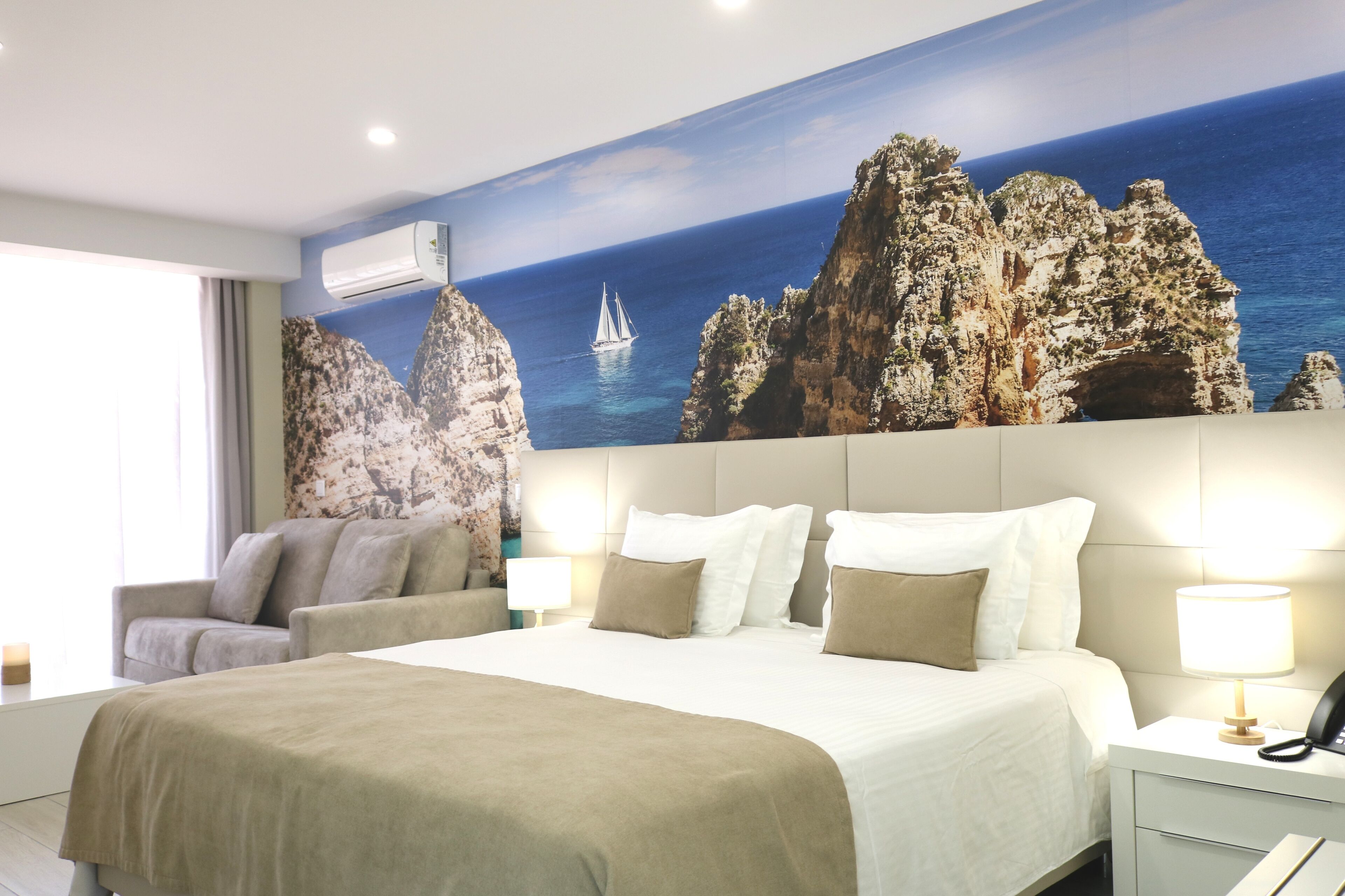 A hotel room with a large bed, a sofa, a nightstand, and a wall mural of a rocky coastline with a sailboat.