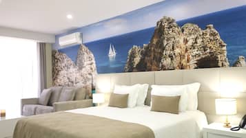 A hotel room with a large bed, a sofa, a nightstand, and a wall mural of a rocky coastline with a sailboat.