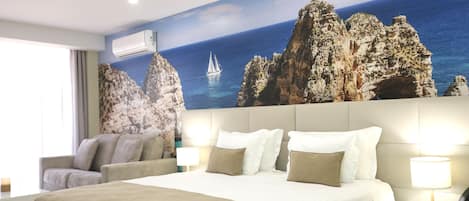 Habitación de hotel con una cama grande, un sofá, una mesita de noche y un mural en la pared que muestra una costa rocosa con un velero.