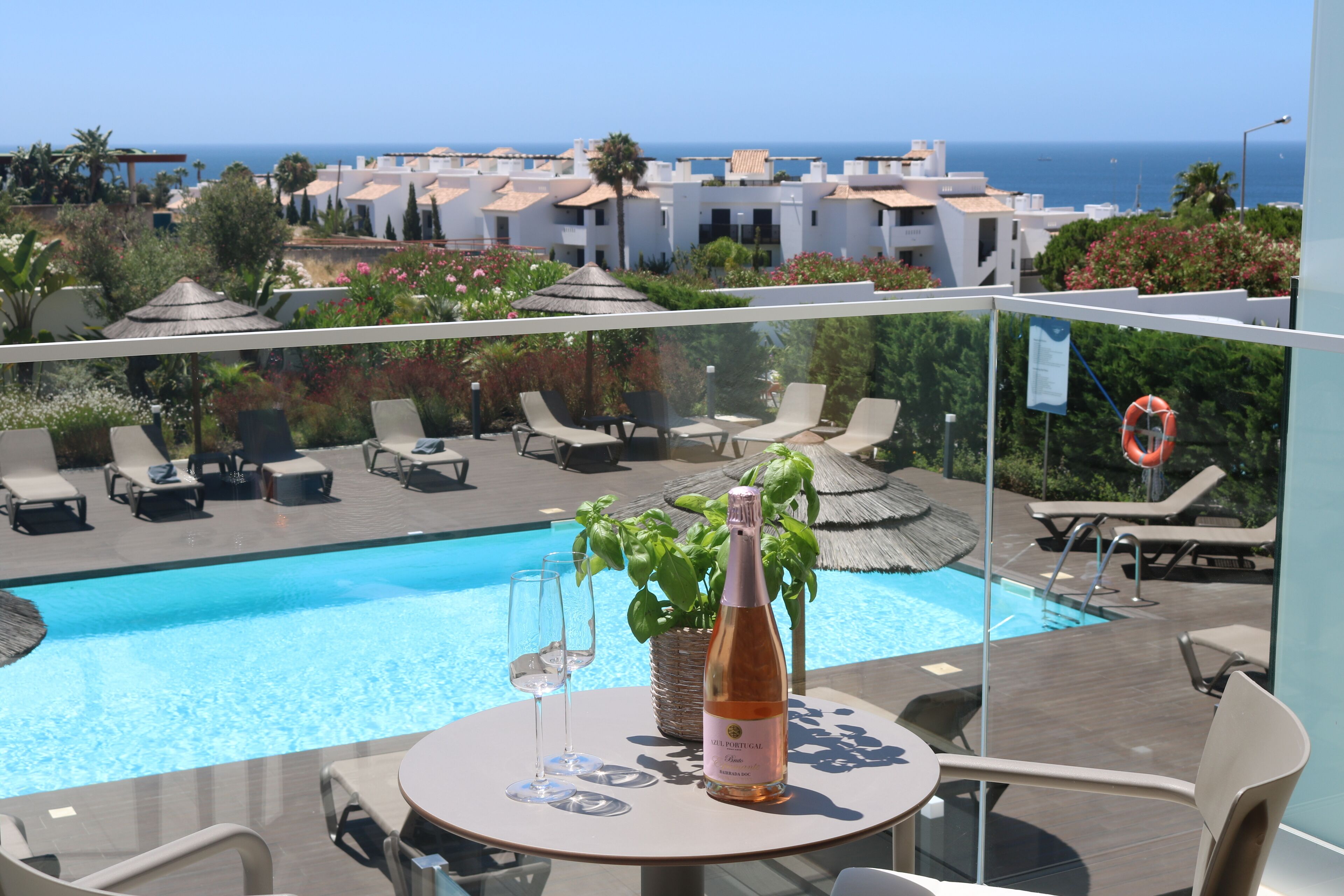 Un área de piscina en la azotea con sillones, una mesa con una botella de vino y vistas al mar y edificios al fondo.