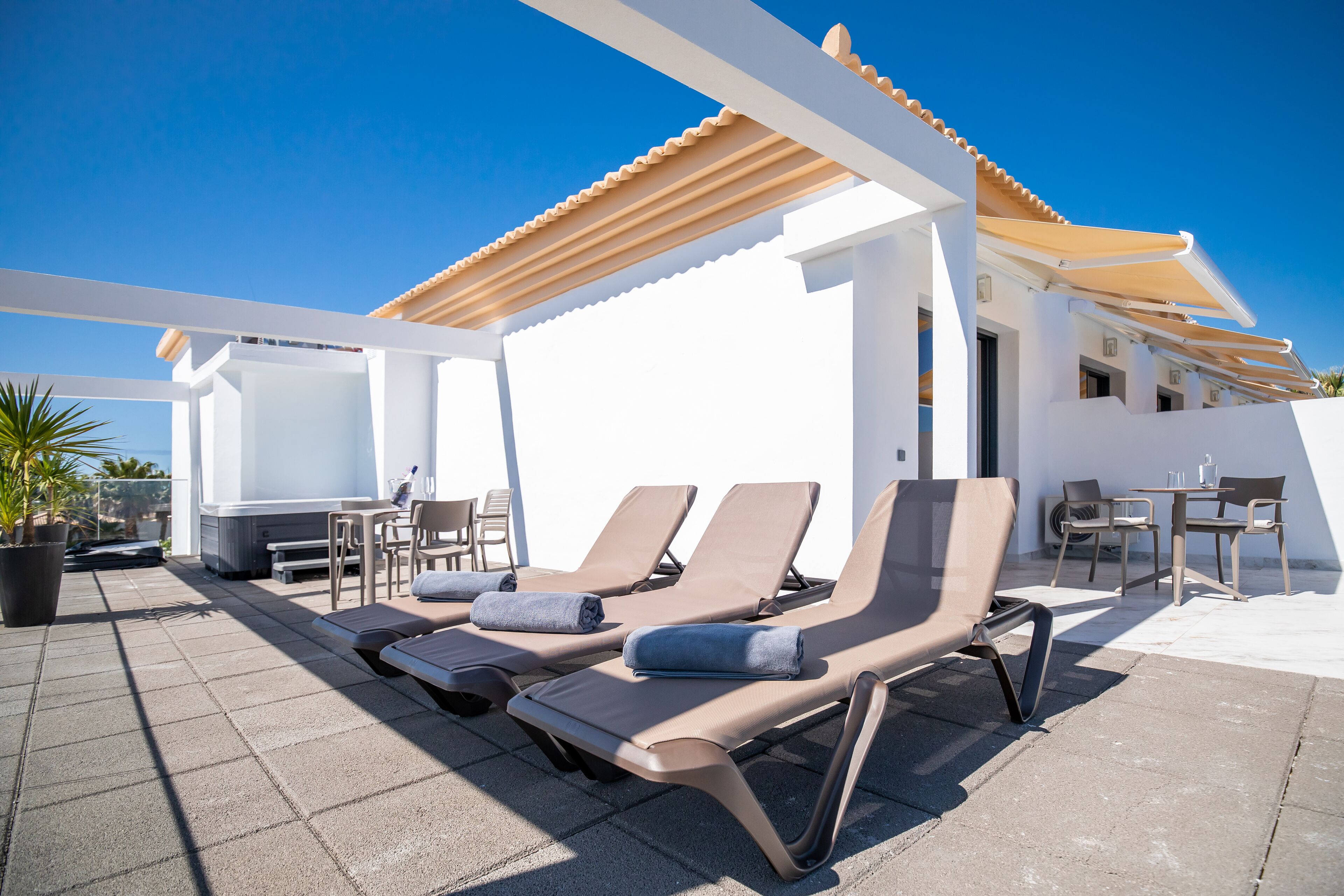A rooftop terrace with sun loungers, a table, chairs, and a potted plant.