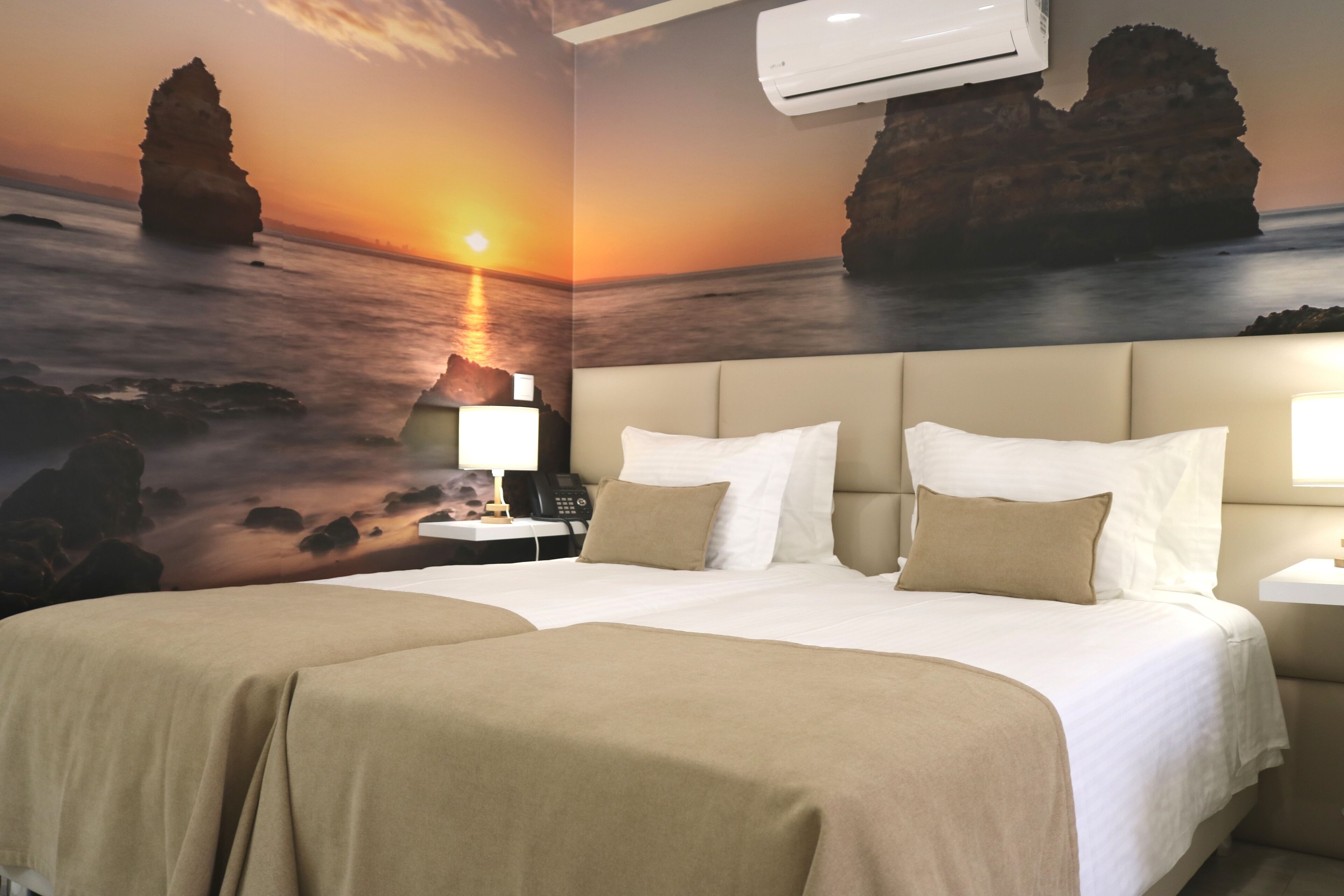A hotel room with a large bed, two bedside tables, a nightstand, and a wall mural of a sunset over the ocean.