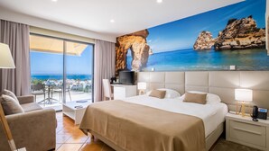 Une chambre d’hôtel avec un grand lit, un bureau, une chaise, une télévision et une vue sur la mer, visible à travers une porte vitrée.