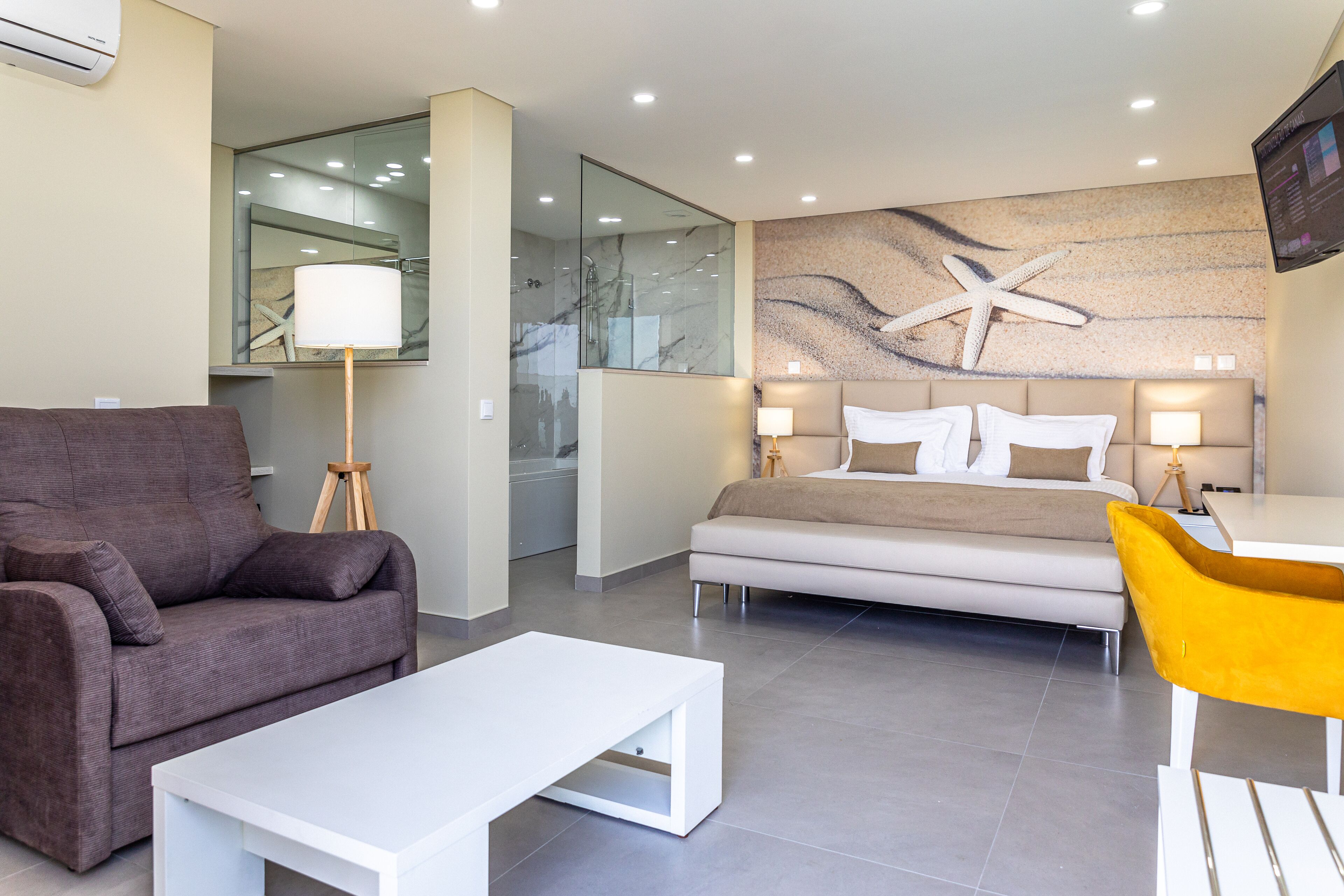A modern hotel room with a sofa, a white coffee table, a yellow chair, a bed with two bedside tables, and a large wall mural.