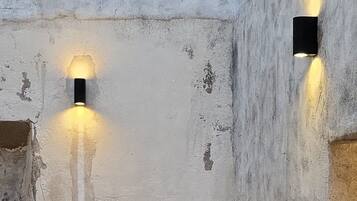 Un interior rústico con una mesa redonda y cuatro sillas de madera, dos lámparas fijadas a la pared y un suelo con diseño de piedra.