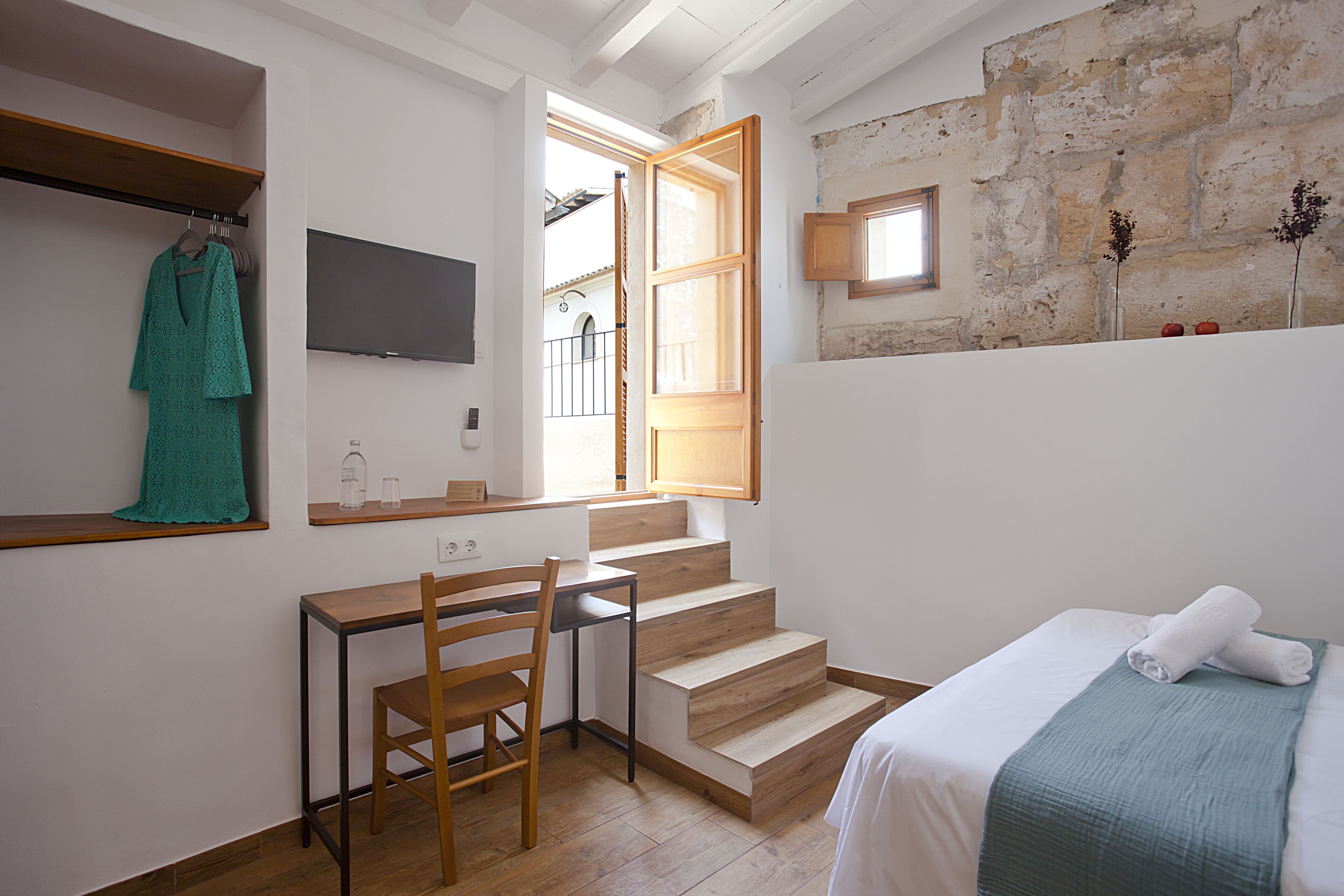 A bedroom with a bed, a desk with a chair, a TV, a window, and a stone wall.
