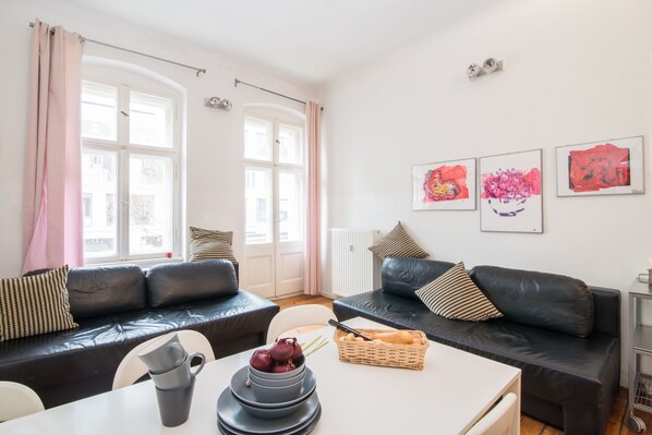 Un salon moderne avec un canapé en cuir noir, une table blanche sur laquelle se trouve un saladier rempli de fruits, et une fenêtre agrémentée de rideaux.