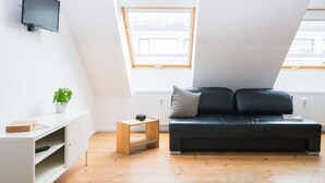 A modern loft bedroom with a sloped ceiling, a black leather sofa, a white TV mounted on the wall, and a small wooden side table with a camera and a plant. - Apartments elPilar Friedrichshain (Berlin)