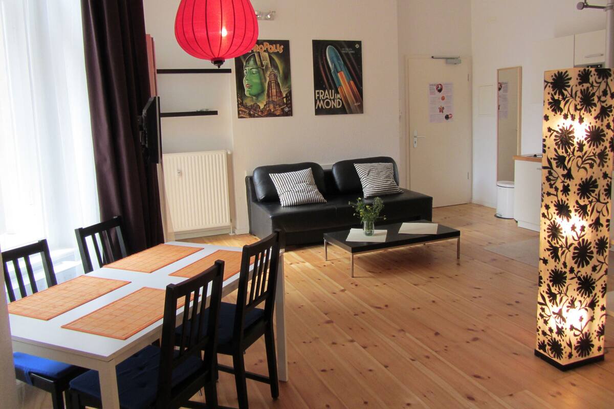 Ein modernes Wohnzimmer mit Essbereich, ausgestattet mit einem schwarzen Sofa, einer roten Pendelleuchte und einer trennenden Wand mit Blumenmuster.