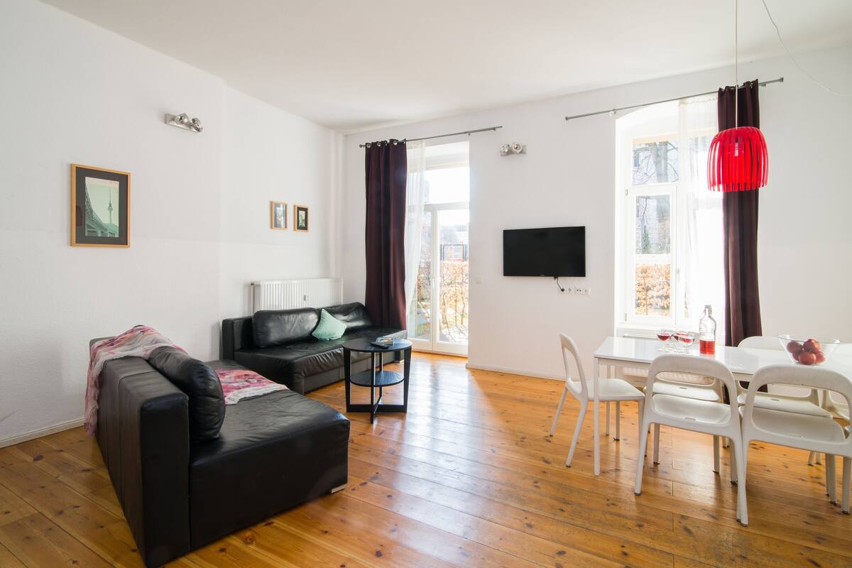 Ein modernes Wohnzimmer mit einer schwarzen Ledersofa, einem kleinen Tisch und einem Essbereich mit weißen Stühlen und einer roten Pendelleuchte.