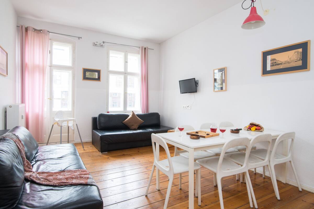 Ein modernes Wohnzimmer mit einer schwarzen Ledersofa, einem weißen Esstisch mit Stühlen, einem an der Wand befestigten Fernseher und einem Fenster mit rosa Vorhängen.