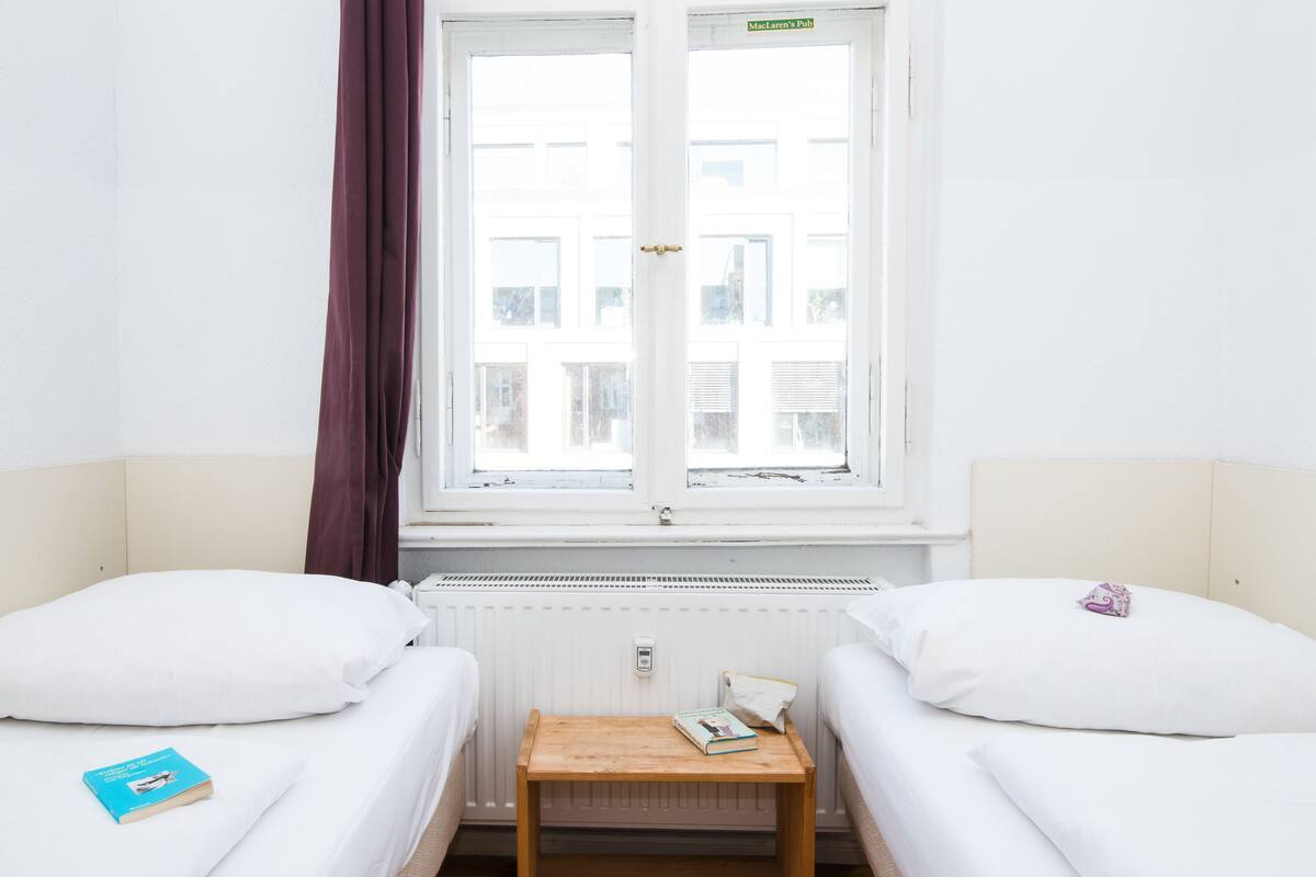 Ein Hotelzimmer mit zwei Betten, einem kleinen Holztisch, einem Fenster mit Blick auf Gebäude und einer Heizung.