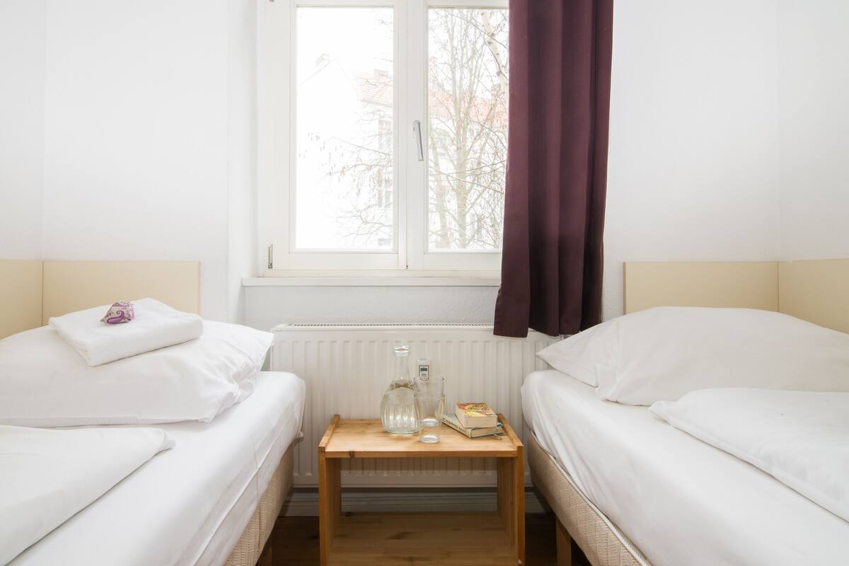 Ein Hotelzimmer mit zwei Betten, einem kleinen Holztisch mit einer Flasche und einem Glas sowie einem Fenster mit Blick auf Bäume.