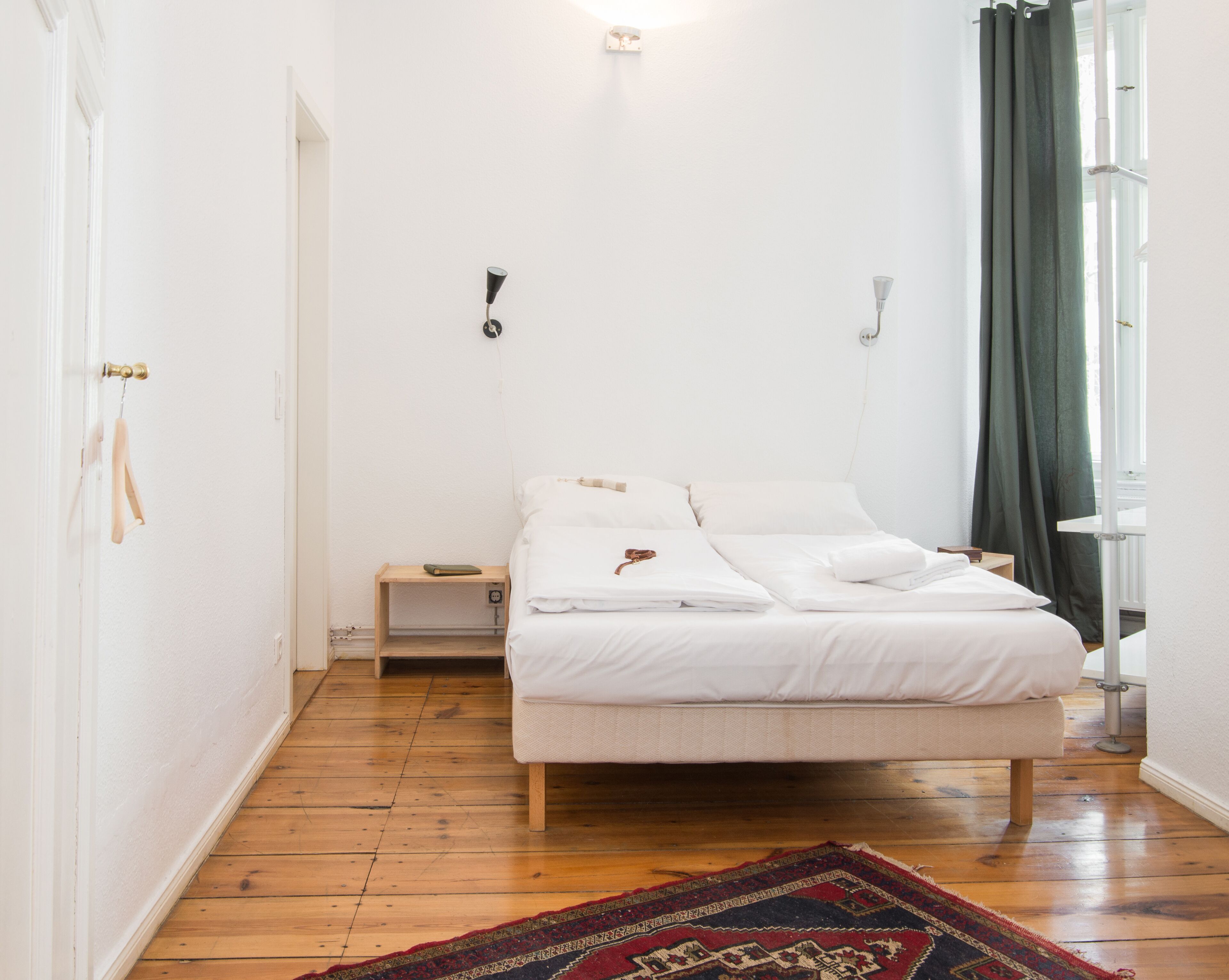 A bedroom with a bed, a bedside table, a nightstand, a wall-mounted lamp, and a window with curtains.