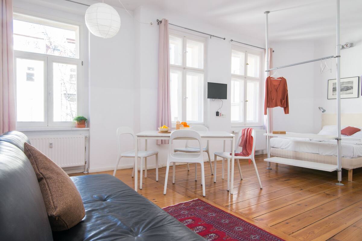 Ein moderner, gut beleuchteter Raum mit einer Couch, einem Esstisch mit Stühlen, einem an der Wand montierten Fernseher und einem Stockbett mit Kleiderhänger.