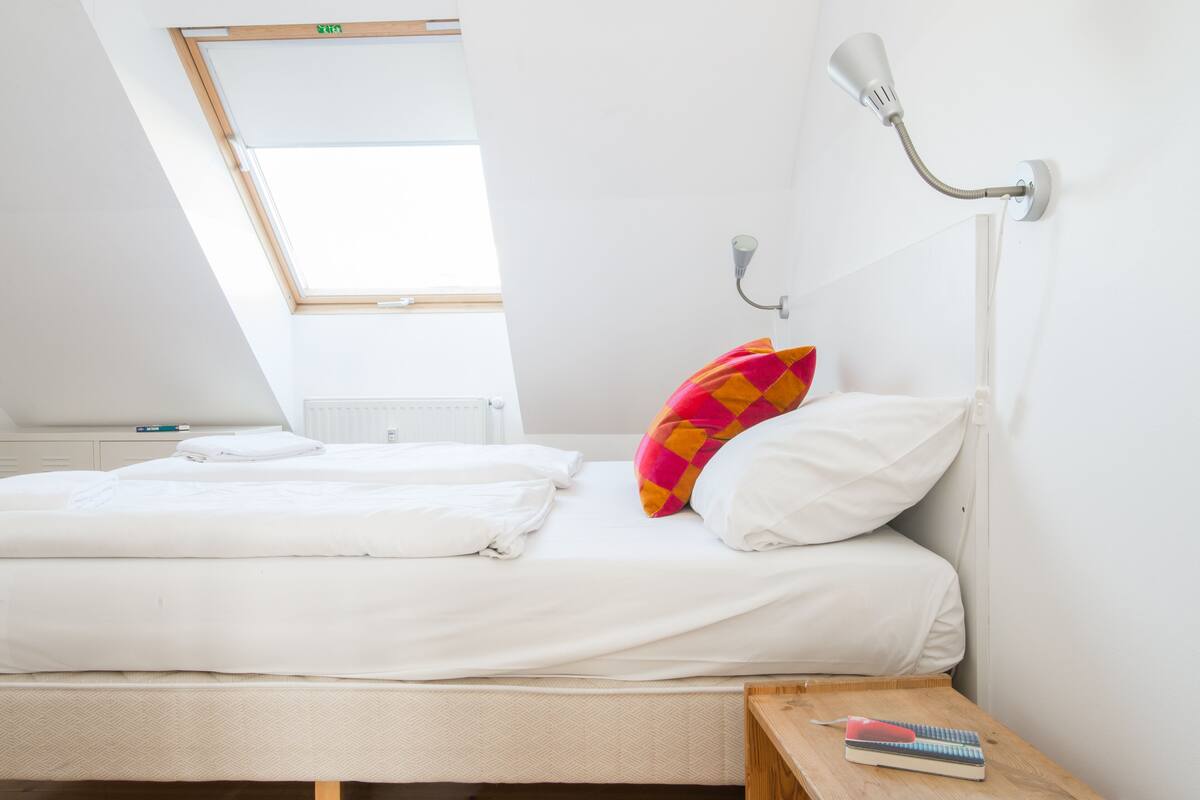 Ein ordentlich bezogenes Doppelbett mit weißer Bettwäsche, ein farbenfrohes Kissen, ein Nachttisch mit Lampe und ein Fenster mit Blick auf den Himmel.