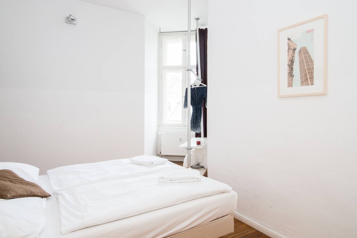 Ein ordentlich bezogenes Bett mit weißen Leinen, einem braunen Kopfkissen und einem weißen Handtuch. An der Wand hängt ein gerahmtes Bild und ein Fenster mit Vorhängen.