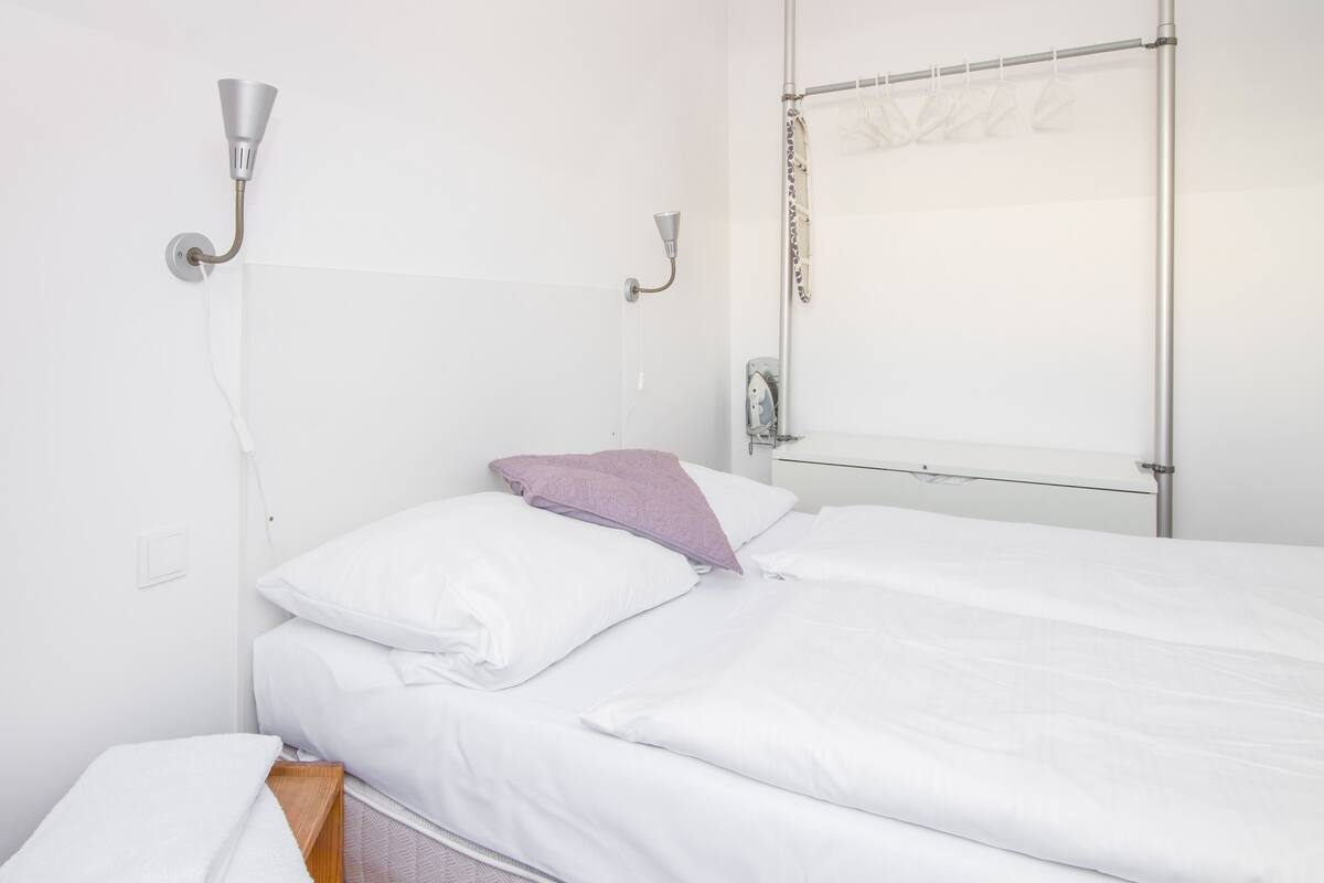 Ein ordentlich bezogenes Bett mit weißen Bettwäsche und einem einzelnen violetten Kissen, eine Wandleuchte und eine Metallvorhangstange mit einem transparenten Vorhang.