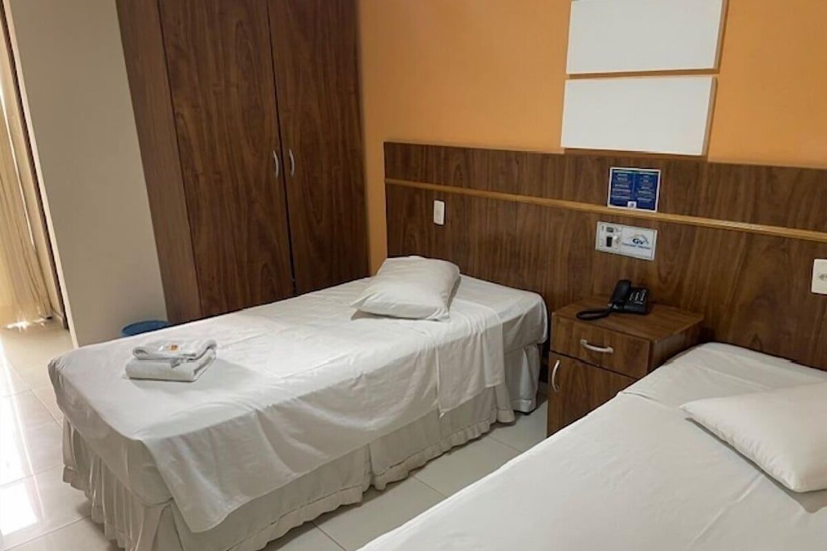 A hotel room with two single beds, a wooden wardrobe, a bedside table with a phone, and a wall-mounted light switch.