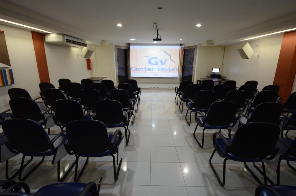 Un salón de conferencias con filas de sillas negras, una pantalla de proyector que muestra 'GV Center Hotel' y un escritorio pequeño con una computadora.