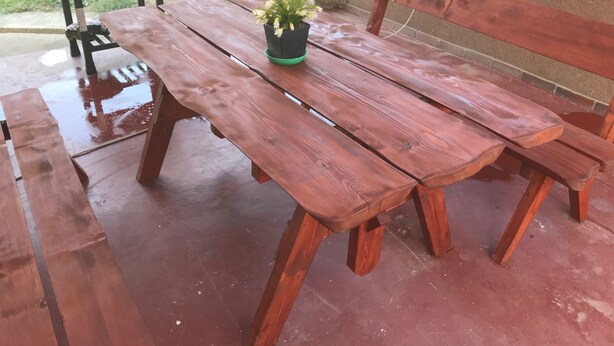 Una taula de picnic de fusta amb una cadira, una petita planta en test i una barbacoa al fons.