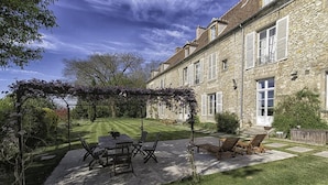 A stone building with a garden patio featuring a pergola, a table with chairs, and a seating area with lounge chairs. - Chambres d'Hôtes de Parseval (Senlis)