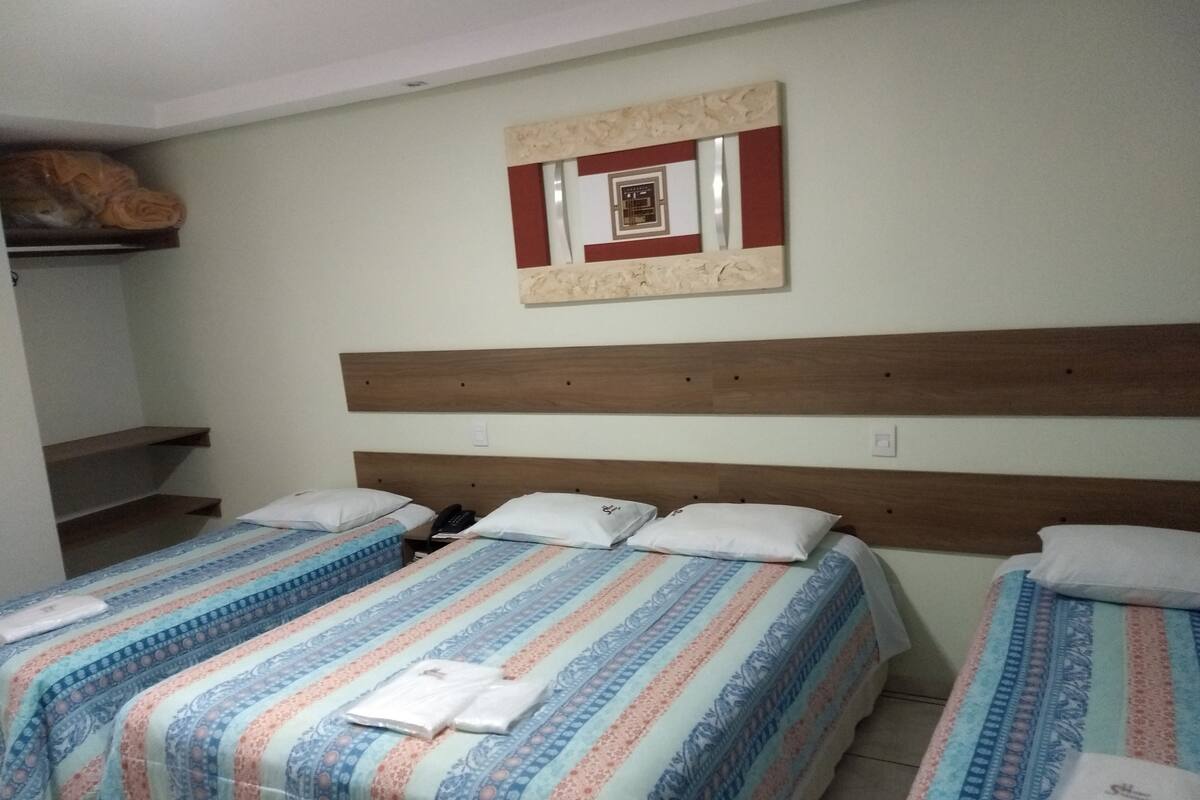 A hotel room with two beds, a wooden headboard, a wall-mounted decoration, and a shelf with a rolled-up item.