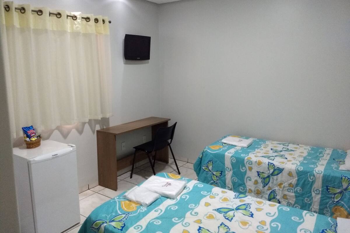 A small, clean room with two beds, a desk, a chair, a TV, and a small refrigerator.