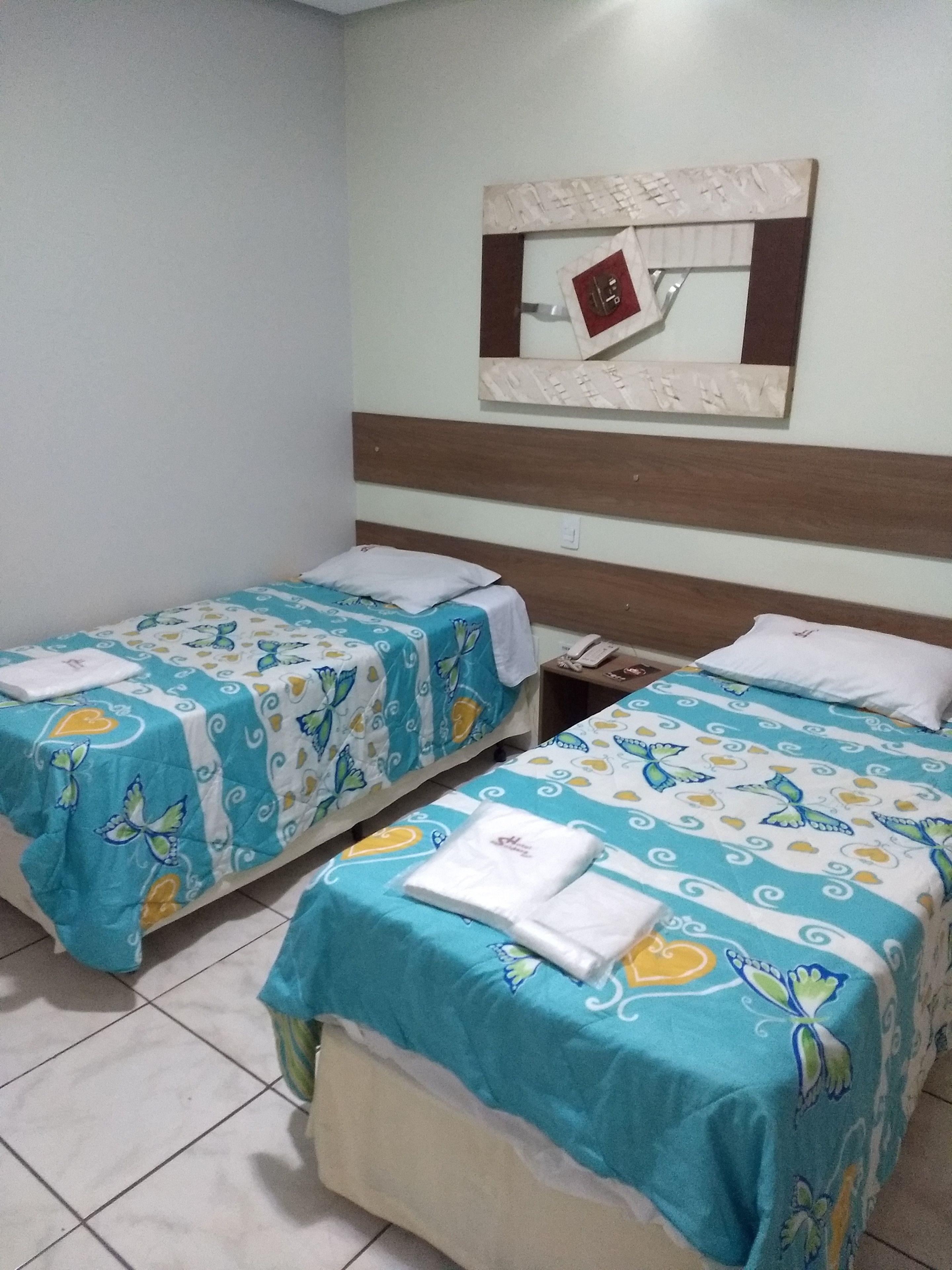 Quarto de hotel com duas camas de solteiro, cada uma com colcha decorada com tema de borboleta em azul e branco. Possui cabeceira de madeira, espelho fixado na parede e pequena mesa de cabeceira com telefone.