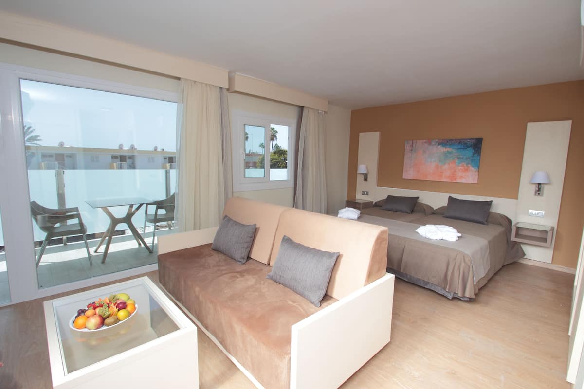 Una habitación de hotel con sofá, cama, una mesita de vidrio con fruta y un balcón con mesa y sillas.