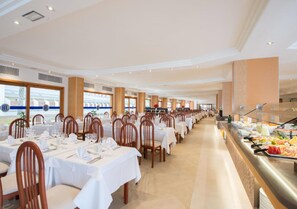 A spacious dining area with multiple tables and chairs, a buffet line, and large windows providing natural light.
