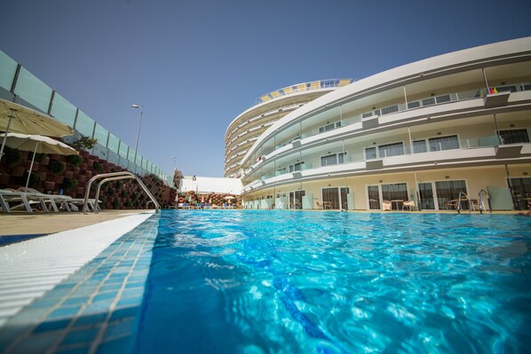 A swimming pool with a clear blue water surface, surrounded by a modern building with multiple balconies and a tiled deck area.