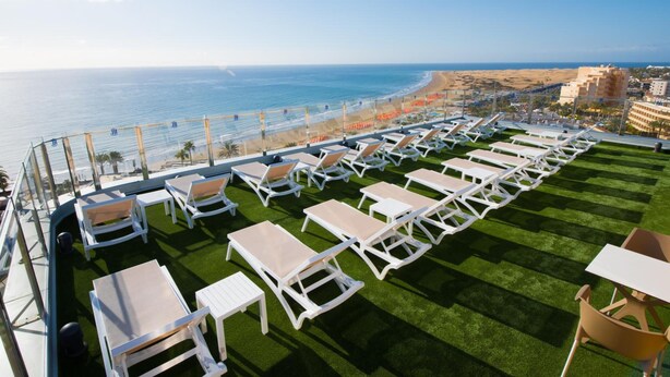 A rooftop lounge with white lounge chairs and tables, overlooking a beach and ocean. - HL Suites Nardos - Only Adults (San Bartolomé de Tirajana)