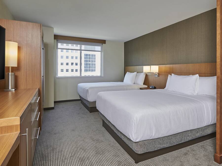 Ein Hotelzimmer mit zwei Betten, einem Schreibtisch, einem Fernseher und einem Fenster mit Blick auf die Stadt.