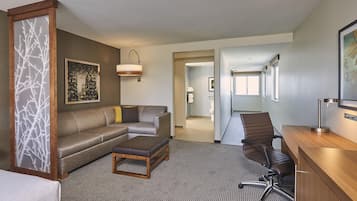 A hotel room with a sofa, a desk, a chair, a coffee table, a bed, a large window, and a cityscape painting.