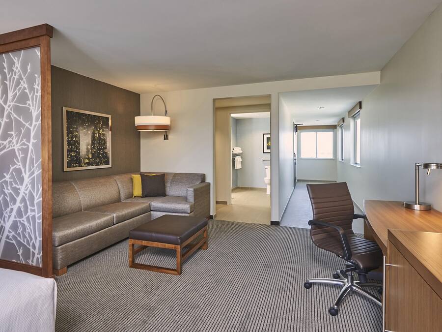 A hotel room with a sofa, a desk, a chair, a coffee table, a bed, a large window, and a cityscape painting.