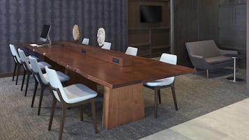 A conference room with a long wooden table, white chairs, a flat-screen TV, and a grey armchair.