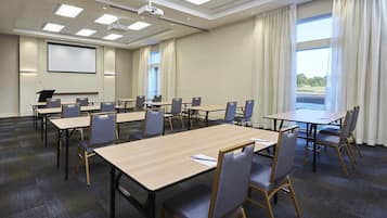 Ein Konferenzraum mit mehreren Tischen und Stühlen, einer Projektionsleinwand und großen Fenstern mit Vorhängen.