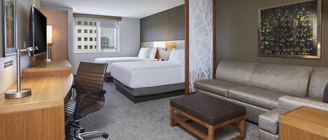 Habitación de hotel con dos camas, un sofá, un escritorio con televisor y vista a la ciudad.