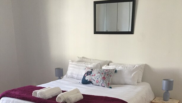 Ein ordentlich bezogenes Bett mit weißen Leinentüchern, einer violetten Tagesdecke, zwei aufgerollten Handtüchern und dekorativen Kissen.