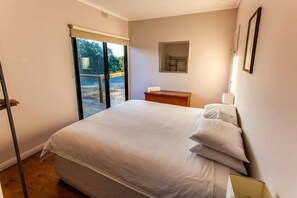 A bedroom with a bed, a small wooden nightstand, a wall-mounted lamp, and a window with a view of the outdoors. - The Kona - coastal seclusion on Kangaroo Island (Penneshaw)