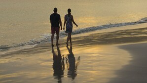 Ein Paar spaziert am Strand entlang, im Hintergrund eine hügelige Landschaft und ruhige See.