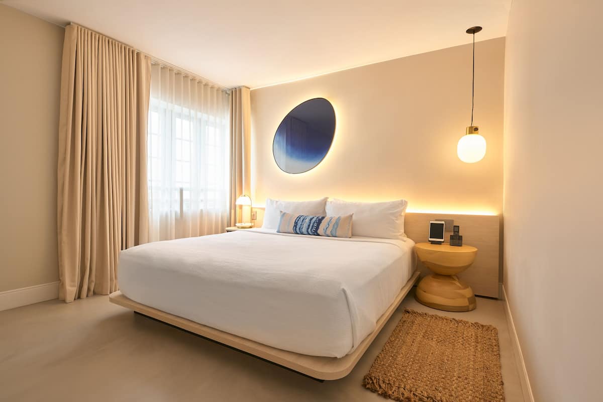 A modern hotel room with a large bed, a bedside table with a phone, and a wall-mounted mirror.