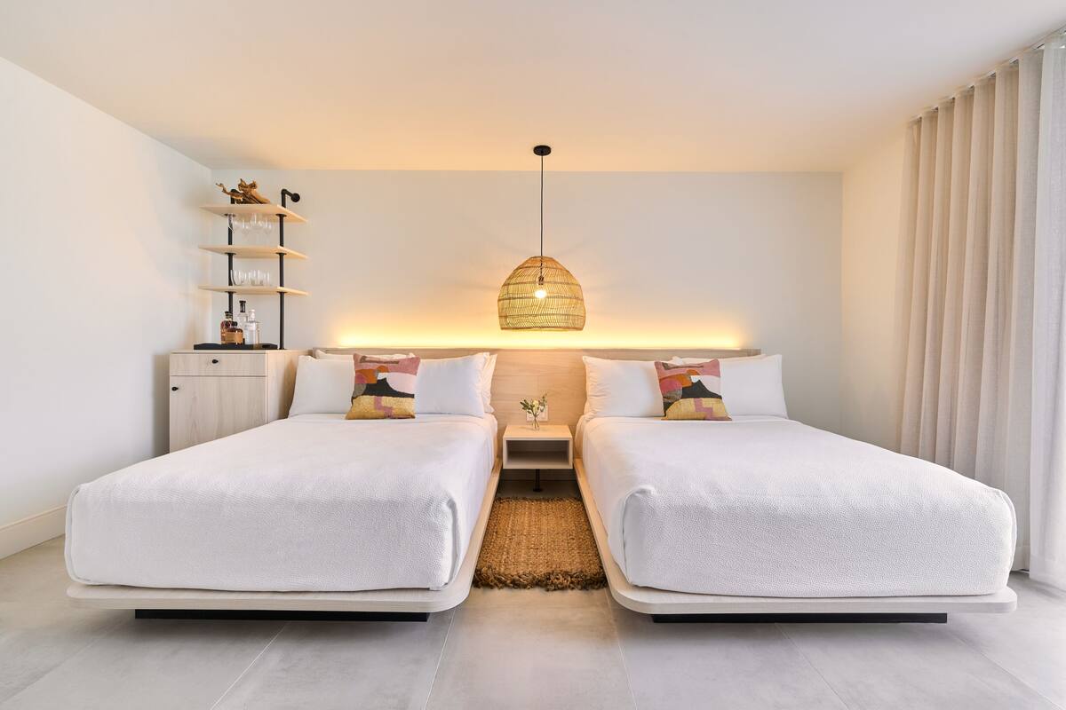 A modern bedroom with two beds, a bedside table, a lamp, and a shelf with decorative items.