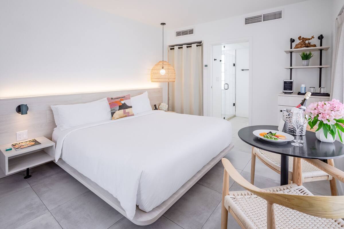 A modern hotel room with a neatly made bed, a bedside table with a lamp, a small table with a vase of flowers, and a chair.