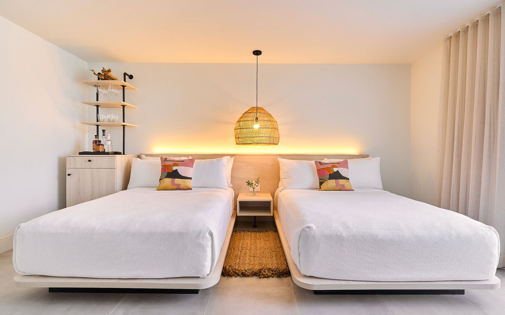 Habitación de hotel con dos camas, una mesita de noche, una lámpara y una estantería con objetos decorativos.