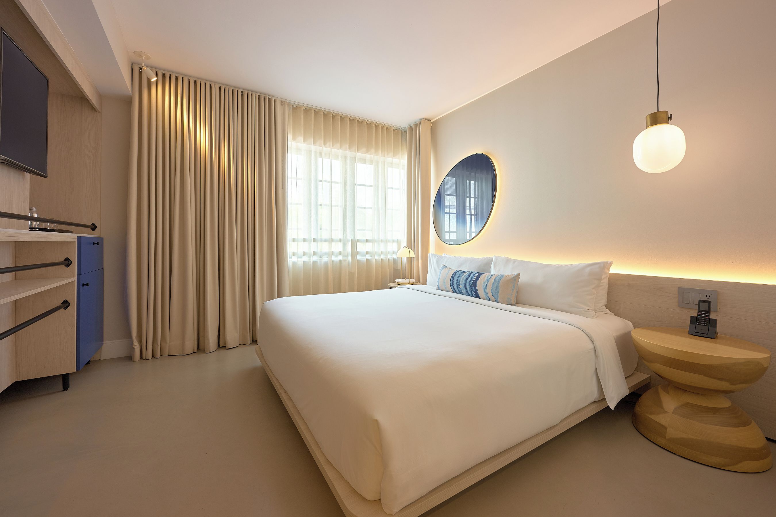 A hotel room with a large bed, a bedside table with a phone, a mirror, and a window with curtains.