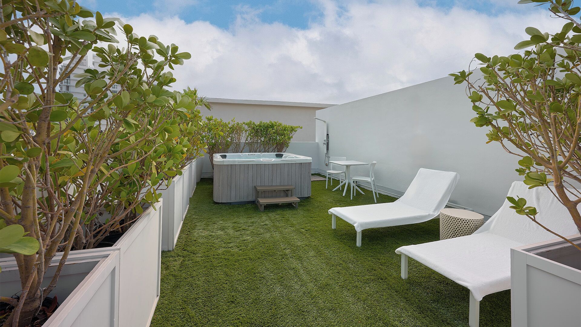 A backyard with artificial grass, a hot tub, white lounge chairs, and potted plants.