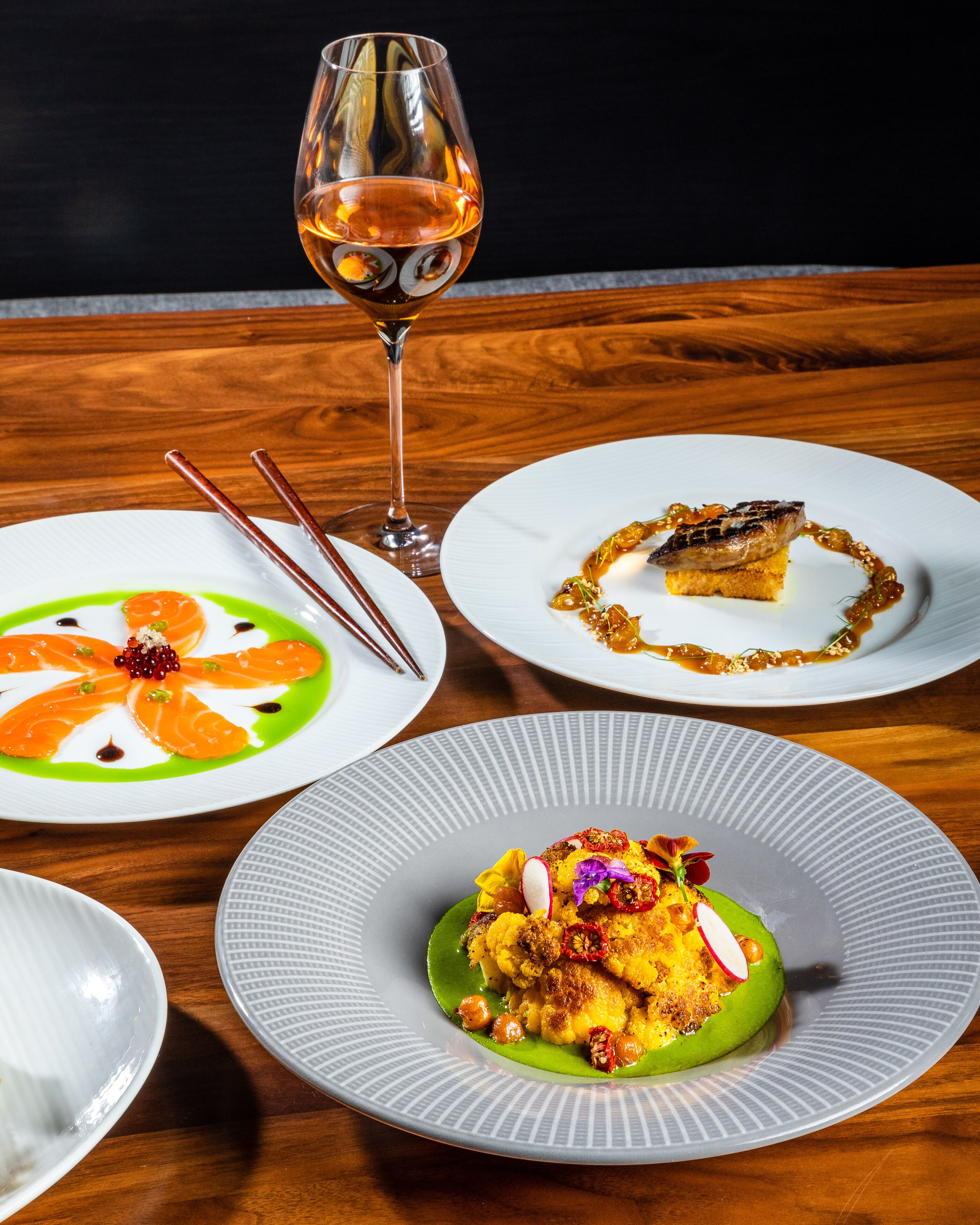 Una mesa con tres platos de comida, una copa de vino y palillos.