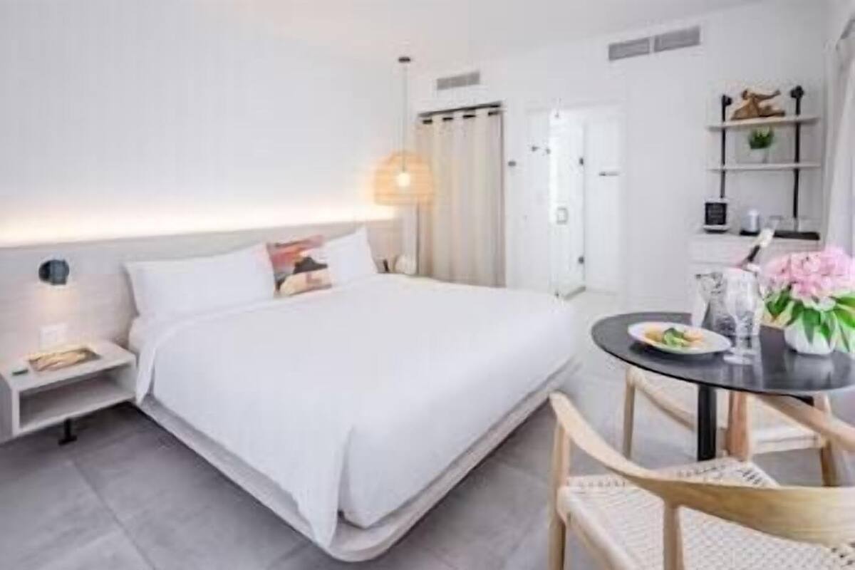 A modern hotel room with a large bed, bedside table, and a small round table with a vase of flowers.